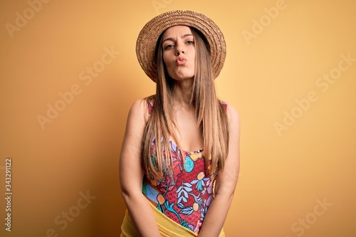 Young beautiful blonde woman wearing swimsuit and summer hat over yellow background looking at the camera blowing a kiss on air being lovely and sexy. Love expression.