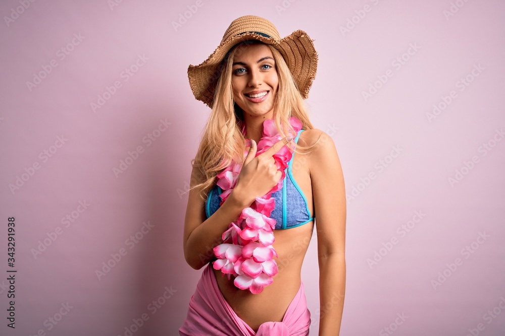 Young beautiful blonde woman on vacation wearing bikini and hat with hawaiian lei flowers cheerful with a smile of face pointing with hand and finger up to the side with happy and natural expression Young beautiful blonde woman on vacation wearing bikini and hat with hawaiian lei flowers cheerful with a smile of face pointing with hand and finger up to the side with happy and natural expression
