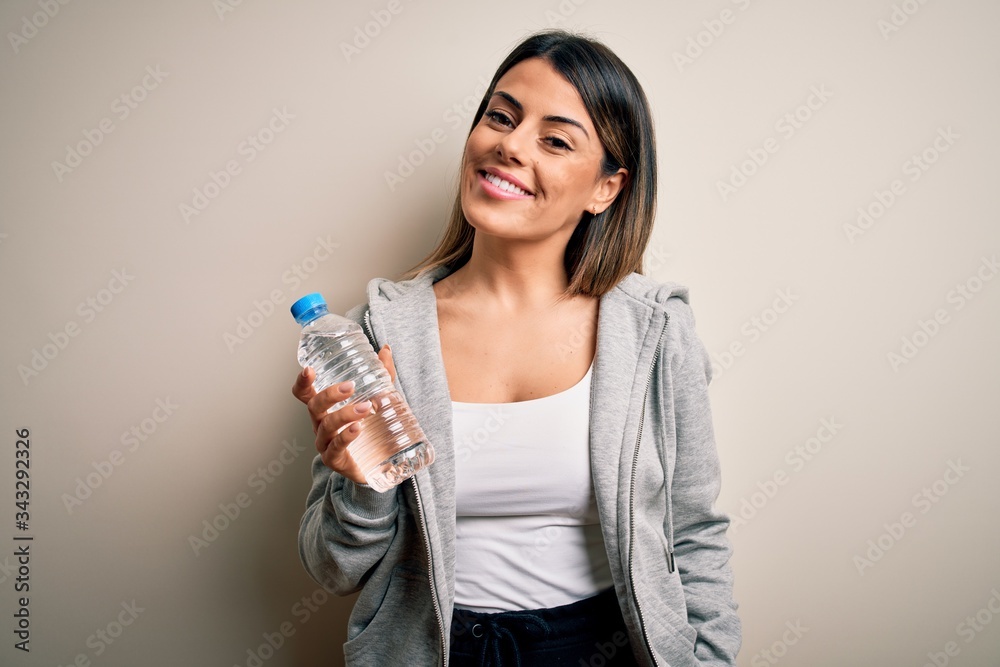 Young beautiful brunette sporty woman drinking bottle of water over isolated white background with a happy face standing and smiling with a confident smile showing teeth Young beautiful brunette sporty woman drinking bottle of water over isolated white background with a happy face standing and smiling with a confident smile showing teeth