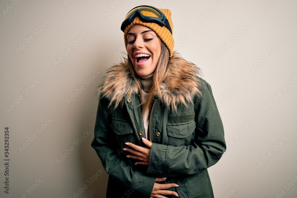 Young brunette skier woman wearing snow clothes and ski goggles over white background smiling and laughing hard out loud because funny crazy joke with hands on body. Young brunette skier woman wearing snow clothes and ski goggles over white background smiling and laughing hard out loud because funny crazy joke with hands on body.