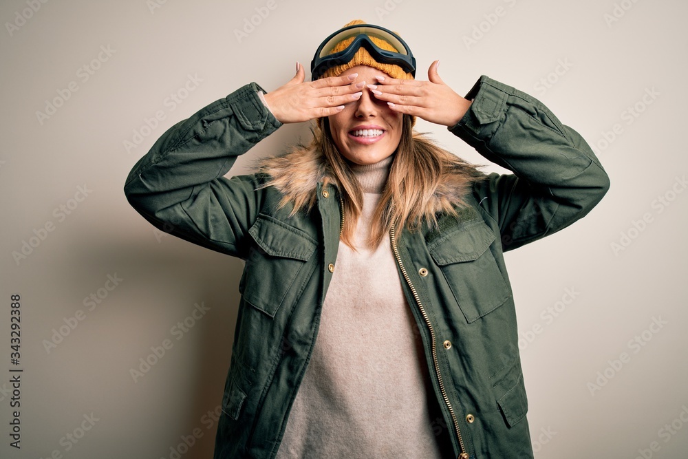 Young brunette skier woman wearing snow clothes and ski goggles over white background covering eyes with hands smiling cheerful and funny. Blind concept. Young brunette skier woman wearing snow clothes and ski goggles over white background covering eyes with hands smiling cheerful and funny. Blind concept.