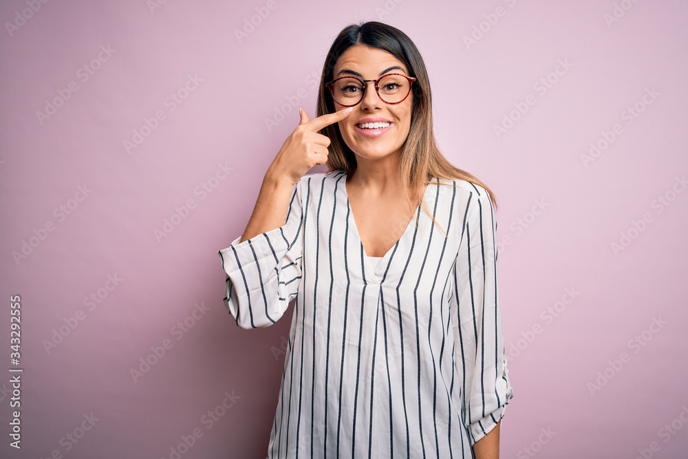 Young beautiful woman wearing casual striped t-shirt and glasses over pink background Pointing with hand finger to face and nose, smiling cheerful. Beauty concept Young beautiful woman wearing casual striped t-shirt and glasses over pink background Pointing with hand finger to face and nose, smiling cheerful. Beauty concept