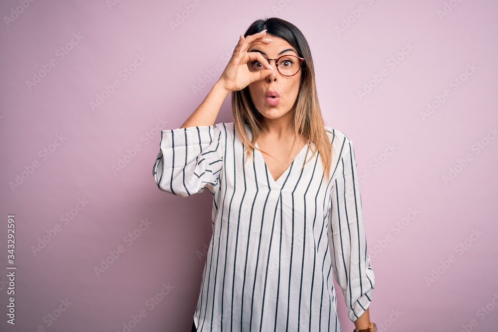 Young beautiful woman wearing casual striped t-shirt and glasses over pink background doing ok gesture shocked with surprised face, eye looking through fingers. Unbelieving expression.