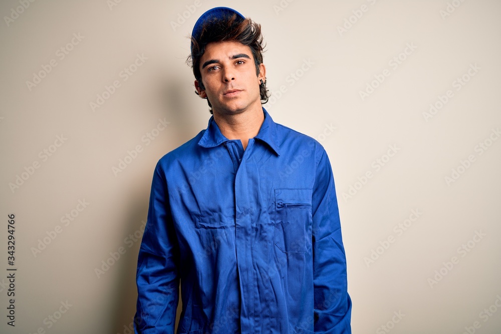 Young mechanic man wearing blue cap and uniform standing over isolated white background Relaxed with serious expression on face. Simple and natural looking at the camera. Young mechanic man wearing blue cap and uniform standing over isolated white background Relaxed with serious expression on face. Simple and natural looking at the camera.