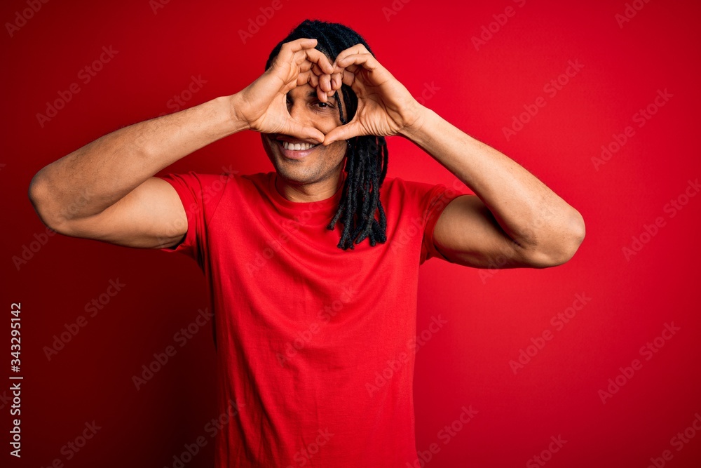 Young handsome african american afro man with dreadlocks wearing red casual t-shirt Doing heart shape with hand and fingers smiling looking through sign