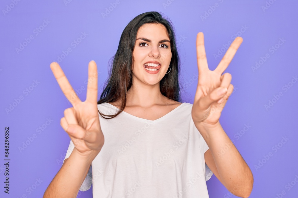 Young beautiful brunette woman wearing casual white t-shirt over purple background smiling with tongue out showing fingers of both hands doing victory sign. Number two. Young beautiful brunette woman wearing casual white t-shirt over purple background smiling with tongue out showing fingers of both hands doing victory sign. Number two.