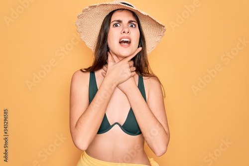 Young beautiful girl wearing swimwear bikini and summer sun hat over yellow background shouting and suffocate because painful strangle. Health problem. Asphyxiate and suicide concept.