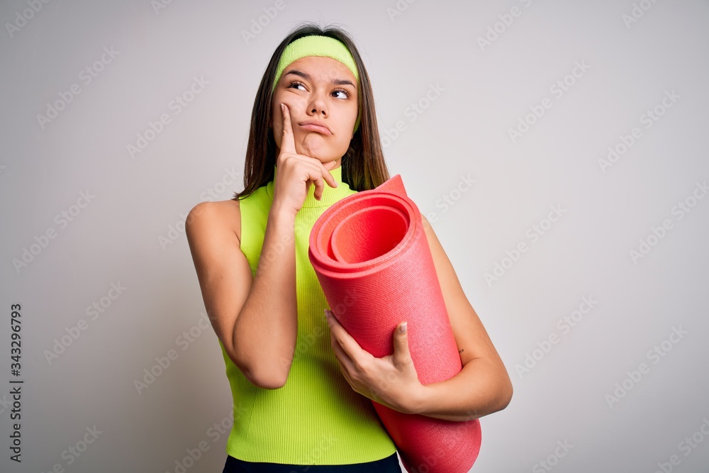 Beautiful sporty girl doing sport wearing sportswear holding yoga mat over white background serious face thinking about question, very confused idea Beautiful sporty girl doing sport wearing sportswear holding yoga mat over white background serious face thinking about question, very confused idea