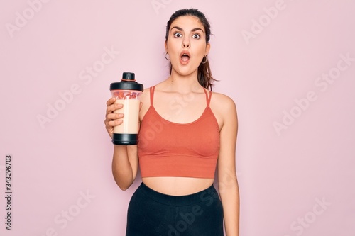 Young beautiful fitness woman wearing sport excersie clothes drinking protein shake scared in shock with a surprise face, afraid and excited with fear expression © Krakenimages.com
