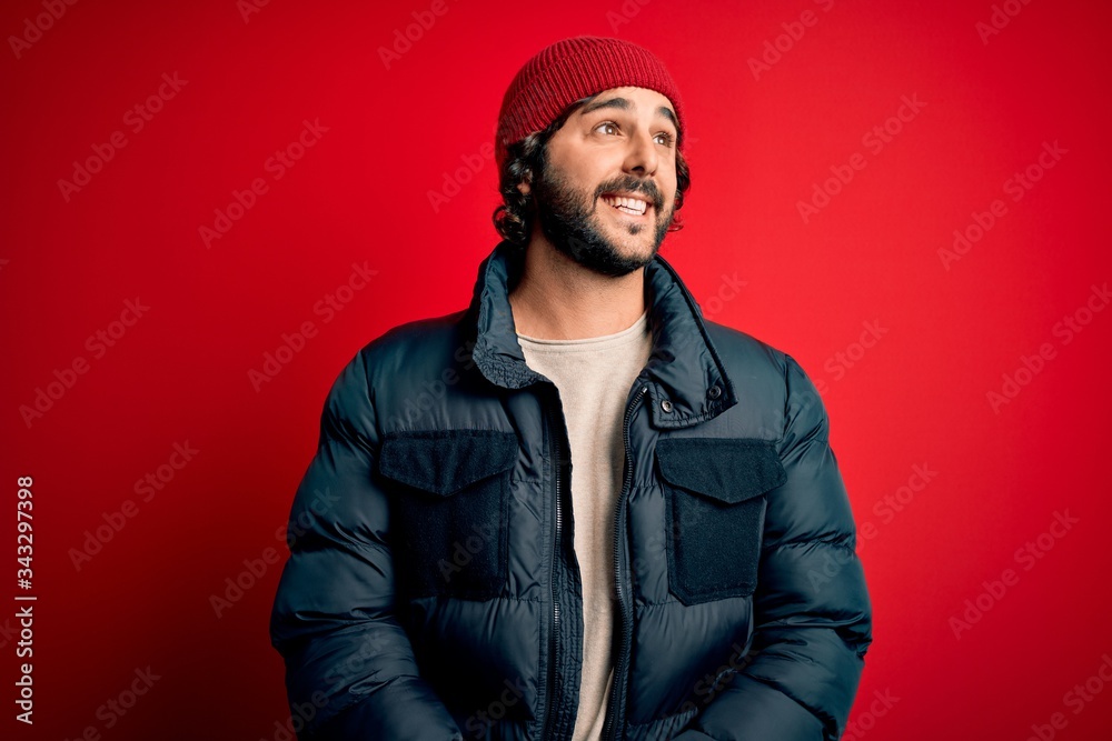 Young handsome man with beard wearing casual coat and wool cap over red background looking away to side with smile on face, natural expression. Laughing confident. Young handsome man with beard wearing casual coat and wool cap over red background looking away to side with smile on face, natural expression. Laughing confident.