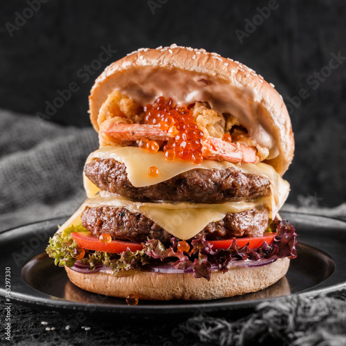Double hamburger or beef burger with red caviar, breaded shrimps, cheese, tomatoes,lettuce and sauce on slate black background, close up