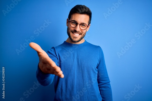 Young handsome man with beard wearing casual sweater and glasses over blue background smiling cheerful offering palm hand giving assistance and acceptance. photo