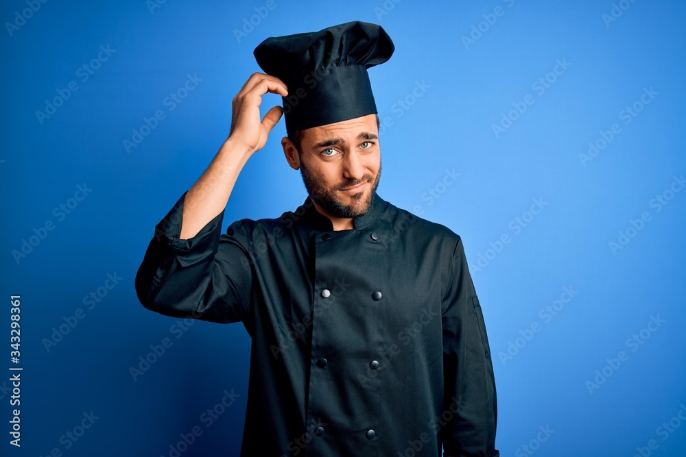 Young handsome chef man with beard wearing cooker uniform and hat over blue background confuse and wonder about question. Uncertain with doubt, thinking with hand on head. Pensive concept. Young handsome chef man with beard wearing cooker uniform and hat over blue background confuse and wonder about question. Uncertain with doubt, thinking with hand on head. Pensive concept.
