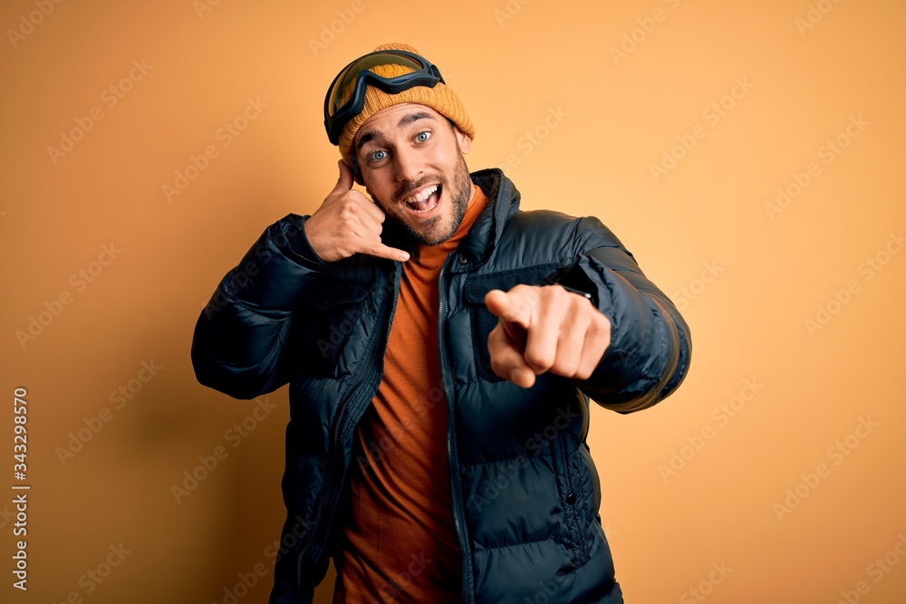 Young handsome skier man with beard wearing snow sportswear and ski goggles smiling doing talking on the telephone gesture and pointing to you. Call me. Young handsome skier man with beard wearing snow sportswear and ski goggles smiling doing talking on the telephone gesture and pointing to you. Call me.