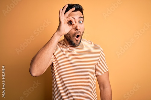 Young handsome man with beard wearing casual striped t-shirt over yellow background doing ok gesture shocked with surprised face, eye looking through fingers. Unbelieving expression.
