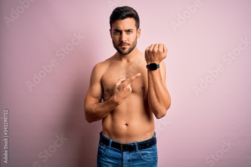 Young handsome strong man with beard shirtless standing over isolated pink background In hurry pointing to watch time, impatience, looking at the camera with relaxed expression