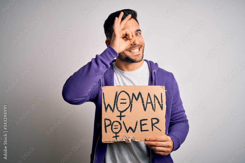 Young handsome activist man protesting for woman power holding cardboard banner with happy face smiling doing ok sign with hand on eye looking through fingers Young handsome activist man protesting for woman power holding cardboard banner with happy face smiling doing ok sign with hand on eye looking through fingers