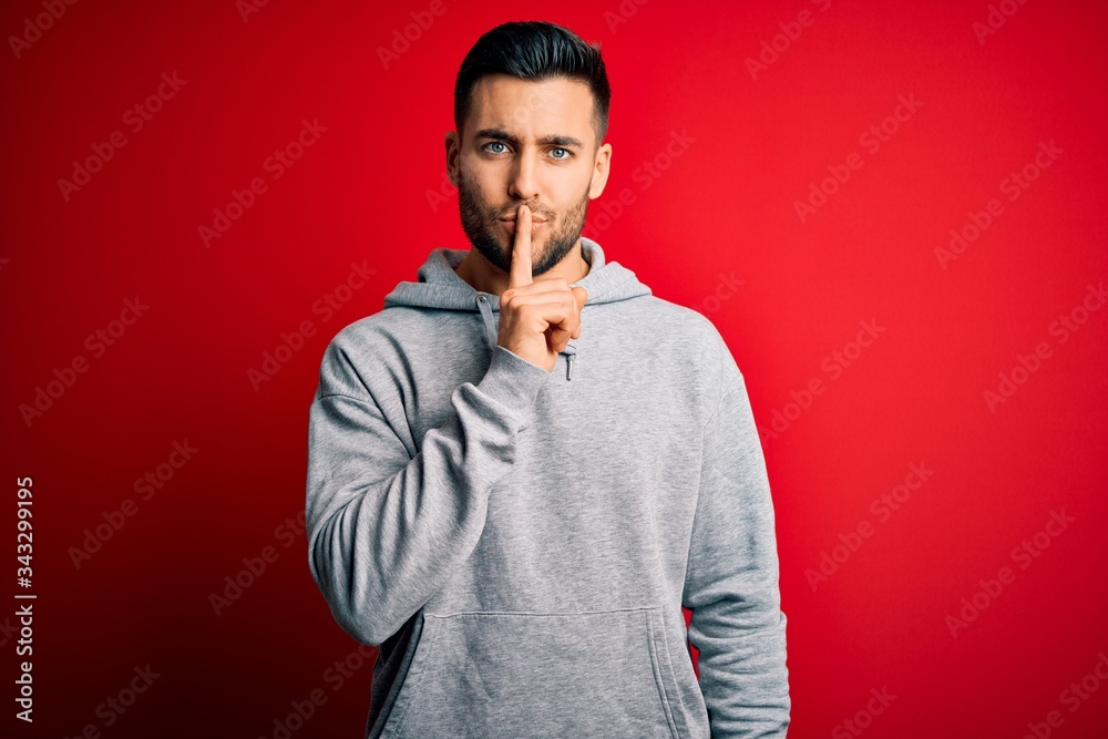 Young handsome sportsman wearing sweatshirt standing over isolated red background asking to be quiet with finger on lips. Silence and secret concept. Young handsome sportsman wearing sweatshirt standing over isolated red background asking to be quiet with finger on lips. Silence and secret concept.