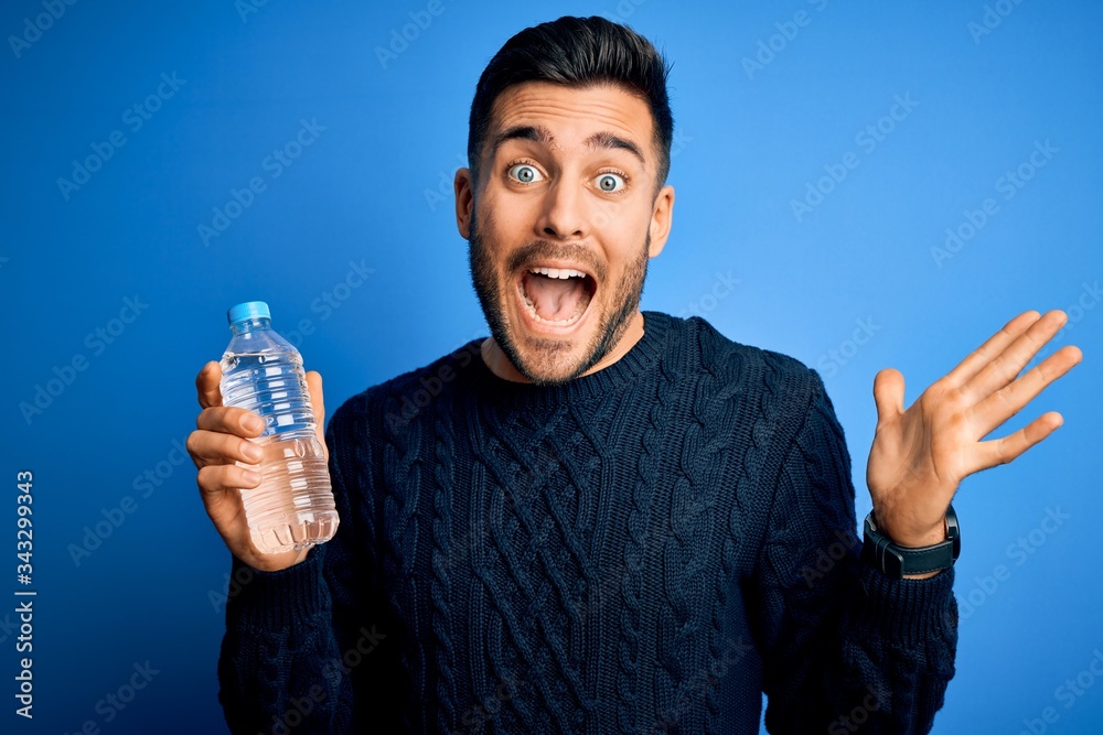Young handsome man drinking bottle of water to refeshment over blue background very happy and excited, winner expression celebrating victory screaming with big smile and raised hands Young handsome man drinking bottle of water to refeshment over blue background very happy and excited, winner expression celebrating victory screaming with big smile and raised hands