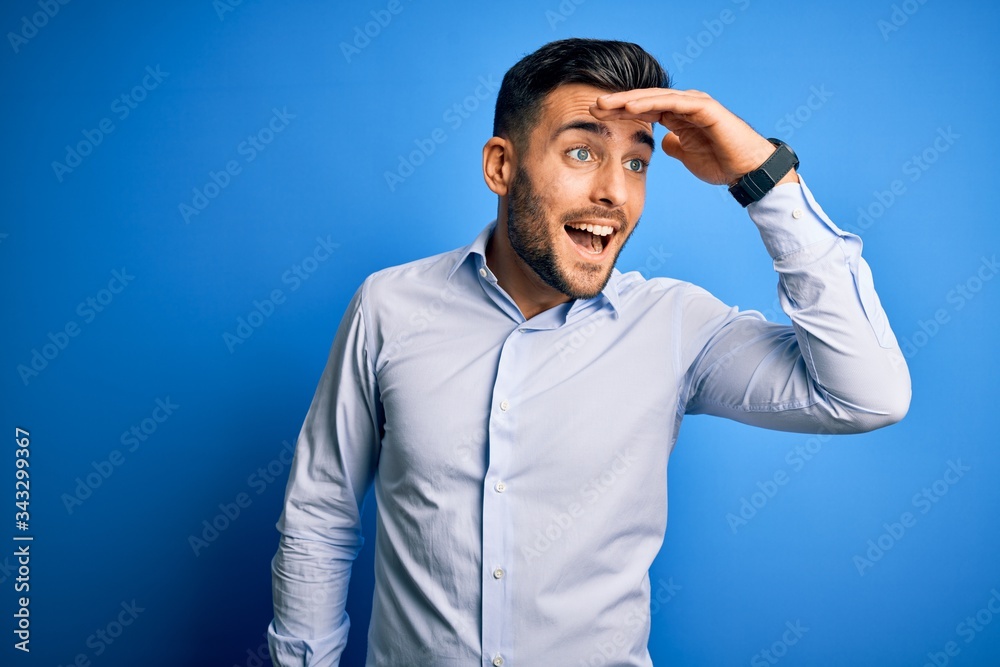 Young handsome man wearing elegant shirt standing over isolated blue background very happy and smiling looking far away with hand over head. Searching concept. Young handsome man wearing elegant shirt standing over isolated blue background very happy and smiling looking far away with hand over head. Searching concept.