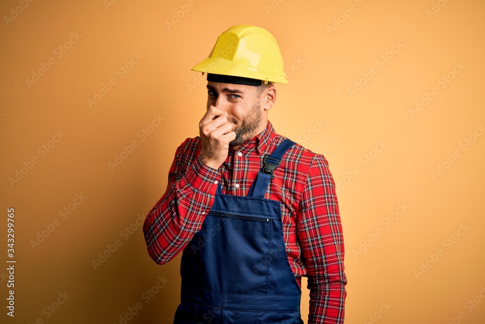Young builder man wearing construction uniform and safety helmet over yellow isolated background smelling something stinky and disgusting, intolerable smell, holding breath with fingers on nose. Bad