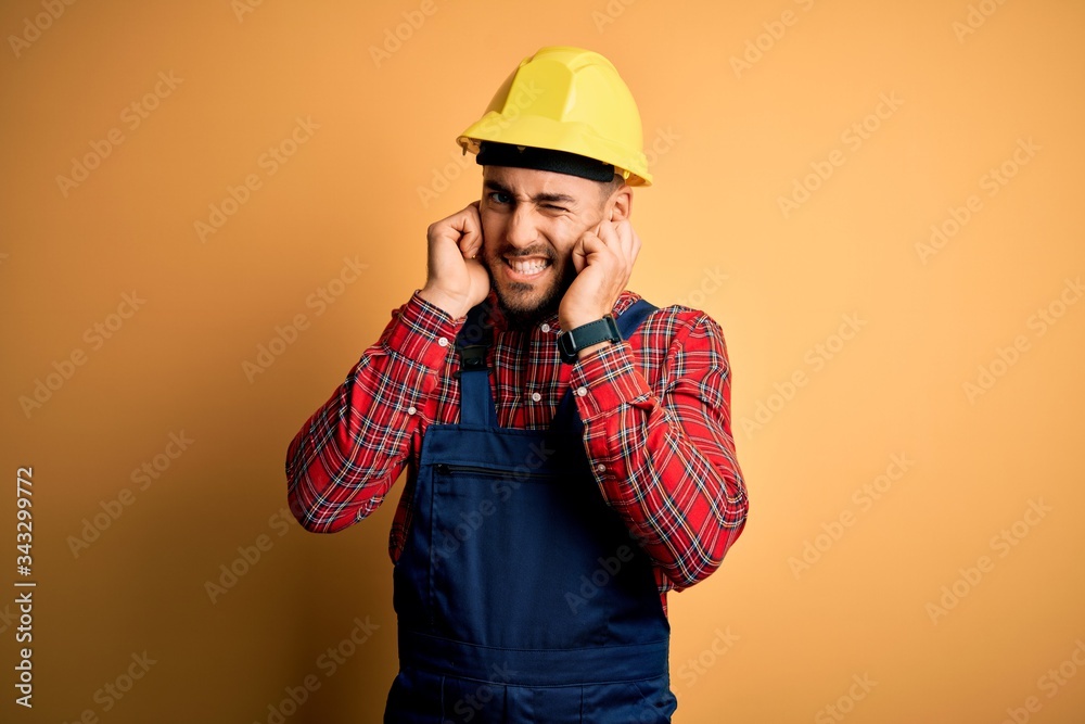 Young builder man wearing construction uniform and safety helmet over yellow isolated background covering ears with fingers with annoyed expression for the noise of loud music. Deaf concept.