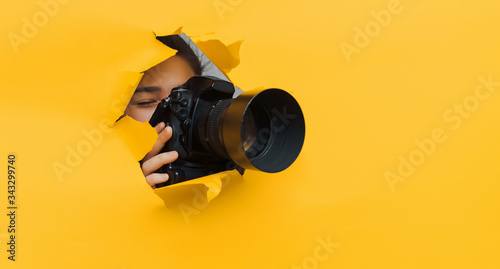 A teenage girl is holding a camera with a telephoto lens that looks out through a torn hole in yellow paper. Concept of paparazzi, espionage, yellow press.