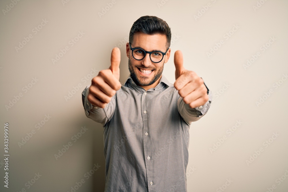 Young handsome man wearing elegant shirt and glasses over isolated white background approving doing positive gesture with hand, thumbs up smiling and happy for success. Winner gesture. Young handsome man wearing elegant shirt and glasses over isolated white background approving doing positive gesture with hand, thumbs up smiling and happy for success. Winner gesture.