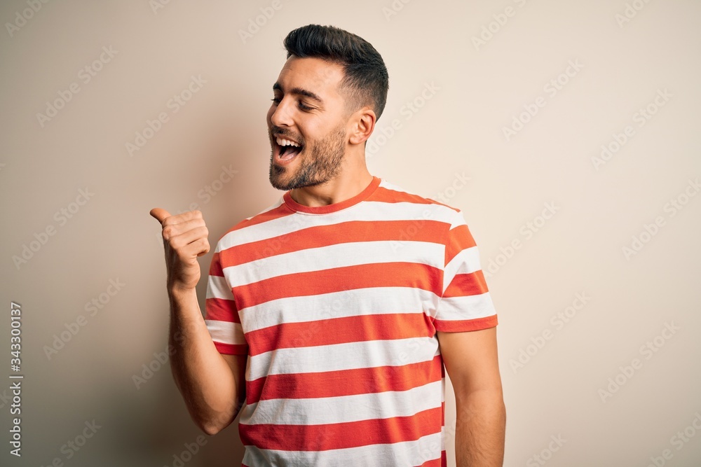 Young handsome man wearing casual striped t-shirt standing over isolated white background smiling with happy face looking and pointing to the side with thumb up. Young handsome man wearing casual striped t-shirt standing over isolated white background smiling with happy face looking and pointing to the side with thumb up.