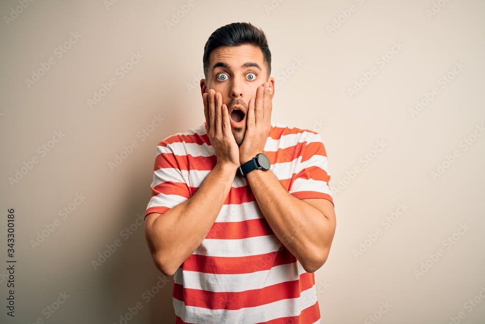 Young handsome man wearing casual striped t-shirt standing over isolated white background afraid and shocked, surprise and amazed expression with hands on face Young handsome man wearing casual striped t-shirt standing over isolated white background afraid and shocked, surprise and amazed expression with hands on face