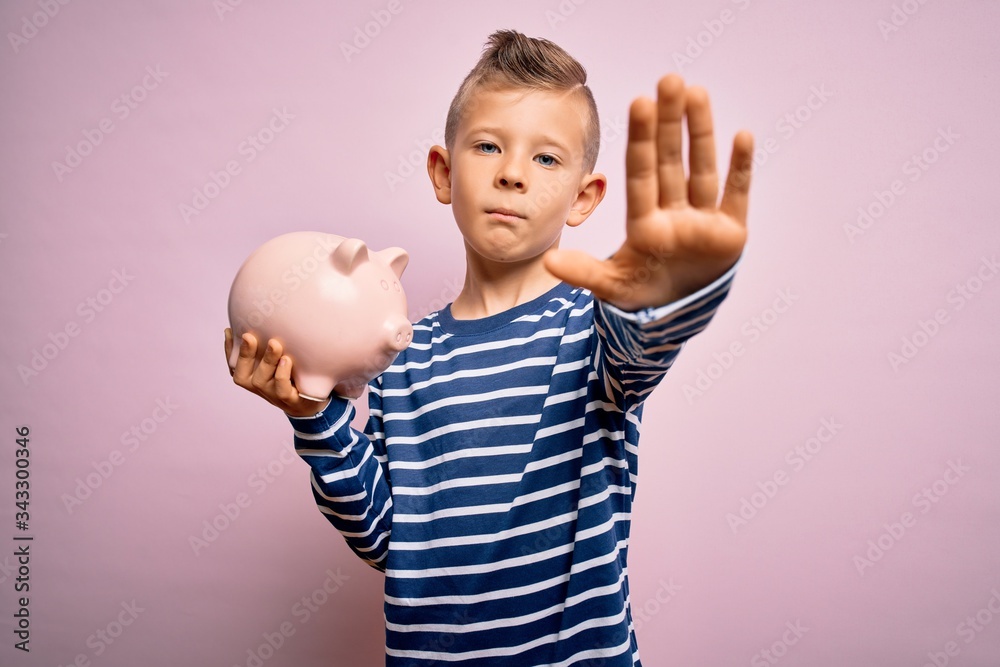 Young little caucasian kid banking money on piggy bank as wealth savings over pink background with open hand doing stop sign with serious and confident expression, defense gesture Young little caucasian kid banking money on piggy bank as wealth savings over pink background with open hand doing stop sign with serious and confident expression, defense gesture