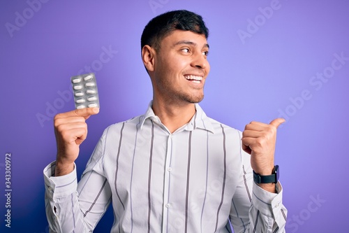 Young hispanic man holding pharmaceutical antibiotics pills over purple isolated background pointing and showing with thumb up to the side with happy face smiling