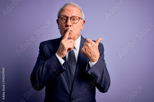 Grey haired senior business man wearing glasses and elegant suit and tie over purple background asking to be quiet with finger on lips pointing with hand to the side. Silence and secret concept. © Krakenimages.com