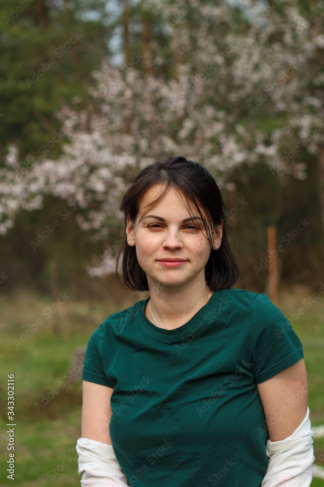 A smiling beautiful, cute white girl with dark hair to the shoulders is standing in a green T-shirt against the background of a blurry flowering spring tree with bokeh. A smiling beautiful, cute white girl with dark hair to the shoulders is standing in a green T-shirt against the background of a blurry flowering spring tree with bokeh.