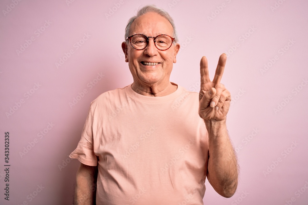 Grey haired senior man wearing glasses standing over pink isolated background smiling with happy face winking at the camera doing victory sign. Number two. Grey haired senior man wearing glasses standing over pink isolated background smiling with happy face winking at the camera doing victory sign. Number two.