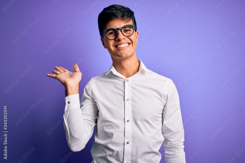 Naklejka premium Young handsome business man wearing shirt and glasses over isolated purple background smiling cheerful presenting and pointing with palm of hand looking at the camera.