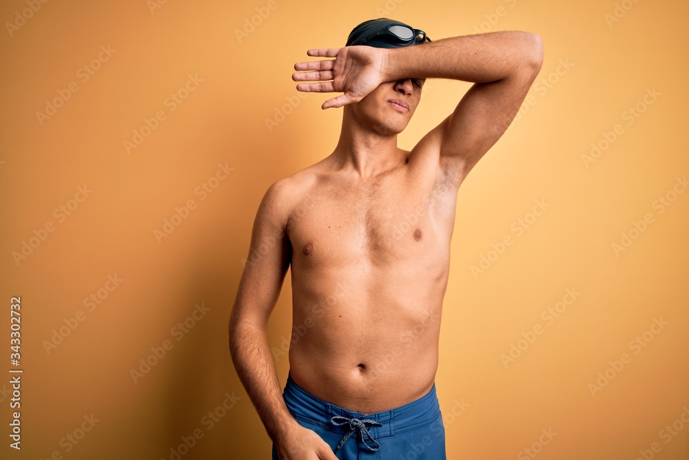 Young handsome man shirtless wearing swimsuit and swim cap over isolated yellow background covering eyes with arm, looking serious and sad. Sightless, hiding and rejection concept