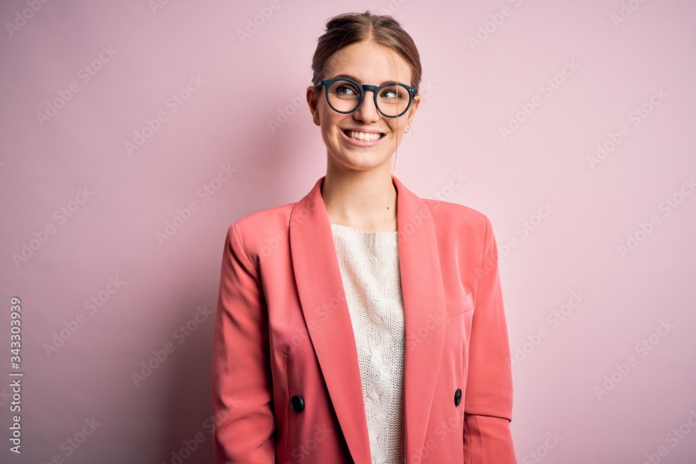 Young beautiful redhead woman wearing jacket and glasses over isolated pink background looking away to side with smile on face, natural expression. Laughing confident.