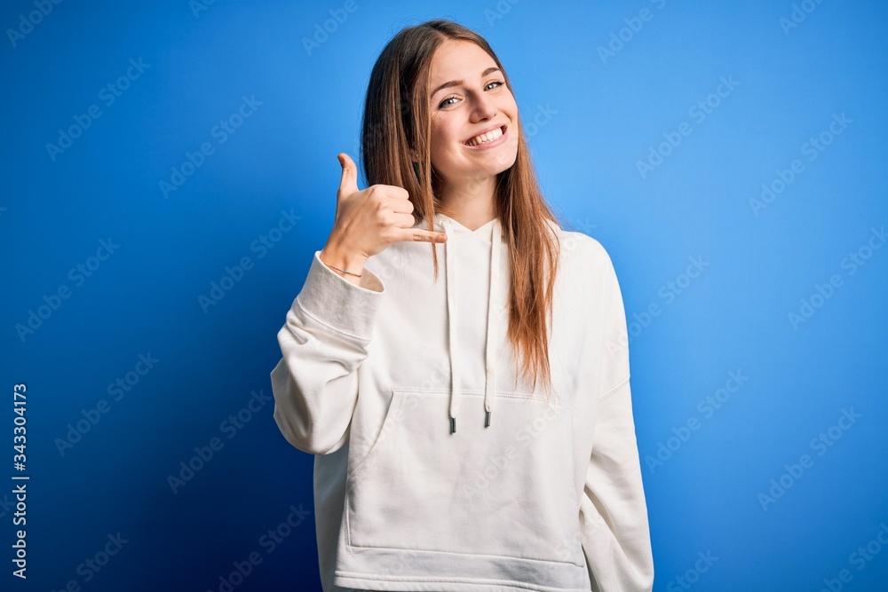 Young beautiful redhead sporty woman wearing sweatshirt over isolated blue background smiling doing phone gesture with hand and fingers like talking on the telephone. Communicating concepts. Young beautiful redhead sporty woman wearing sweatshirt over isolated blue background smiling doing phone gesture with hand and fingers like talking on the telephone. Communicating concepts.
