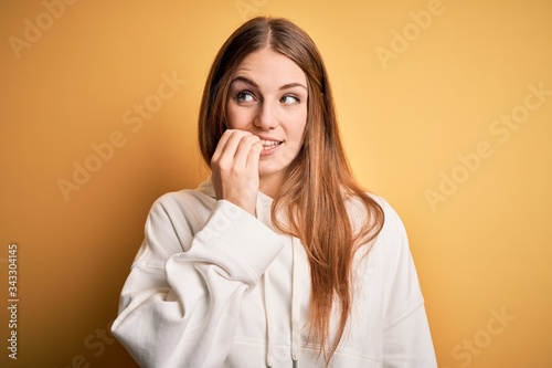 Young beautiful redhead sporty woman wearing sweatshirt over isolated yellow background looking stressed and nervous with hands on mouth biting nails. Anxiety problem.