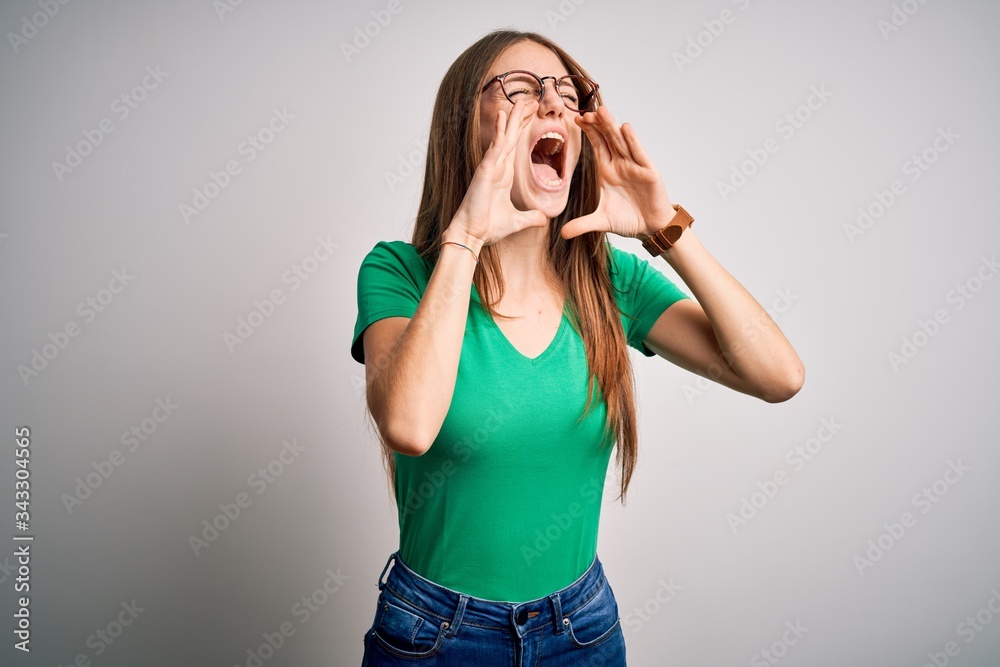 Young beautiful redhead woman wearing casual green t-shirt and glasses over white background Shouting angry out loud with hands over mouth Young beautiful redhead woman wearing casual green t-shirt and glasses over white background Shouting angry out loud with hands over mouth