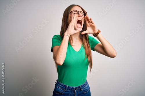 Young beautiful redhead woman wearing casual green t-shirt and glasses over white background Shouting angry out loud with hands over mouth © Krakenimages.com