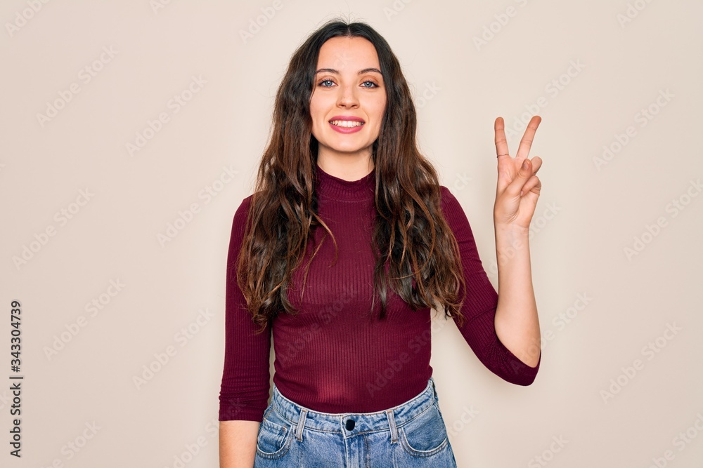 Young beautiful woman wearing casual t-shirt standing over isolated white background smiling with happy face winking at the camera doing victory sign. Number two. Young beautiful woman wearing casual t-shirt standing over isolated white background smiling with happy face winking at the camera doing victory sign. Number two.