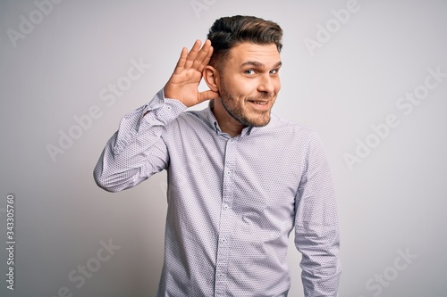 Young business man with blue eyes standing over isolated background smiling with hand over ear listening an hearing to rumor or gossip. Deafness concept. © Krakenimages.com