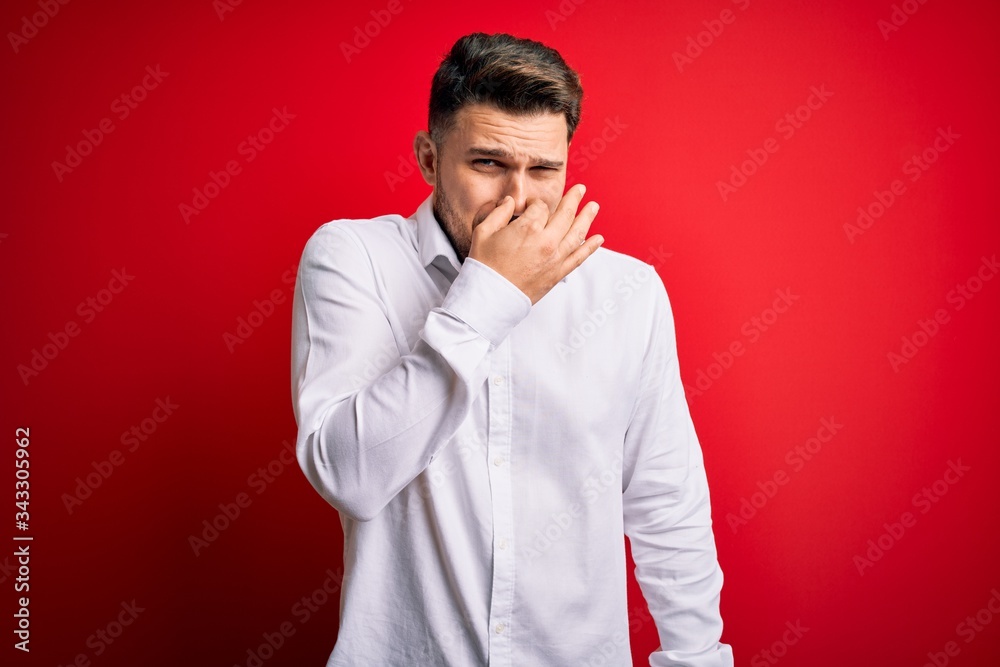 Young business man with blue eyes wearing elegant shirt standing over red isolated background smelling something stinky and disgusting, intolerable smell, holding breath with fingers on the nose. Bad Young business man with blue eyes wearing elegant shirt standing over red isolated background smelling something stinky and disgusting, intolerable smell, holding breath with fingers on the nose. Bad