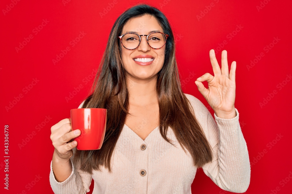 Young hispanic woman wearing glasses drinking a cup of coffee over red background doing ok sign with fingers, excellent symbol Young hispanic woman wearing glasses drinking a cup of coffee over red background doing ok sign with fingers, excellent symbol