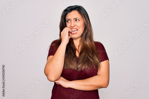 Young hispanic woman wearing casual t-shirt standing over isolated background looking stressed and nervous with hands on mouth biting nails. Anxiety problem.