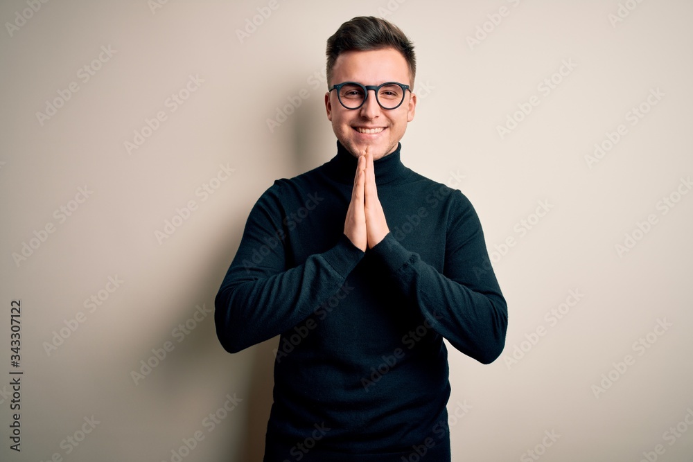 Young handsome caucasian man wearing glasses and casual sweater over isolated background praying with hands together asking for forgiveness smiling confident. Young handsome caucasian man wearing glasses and casual sweater over isolated background praying with hands together asking for forgiveness smiling confident.