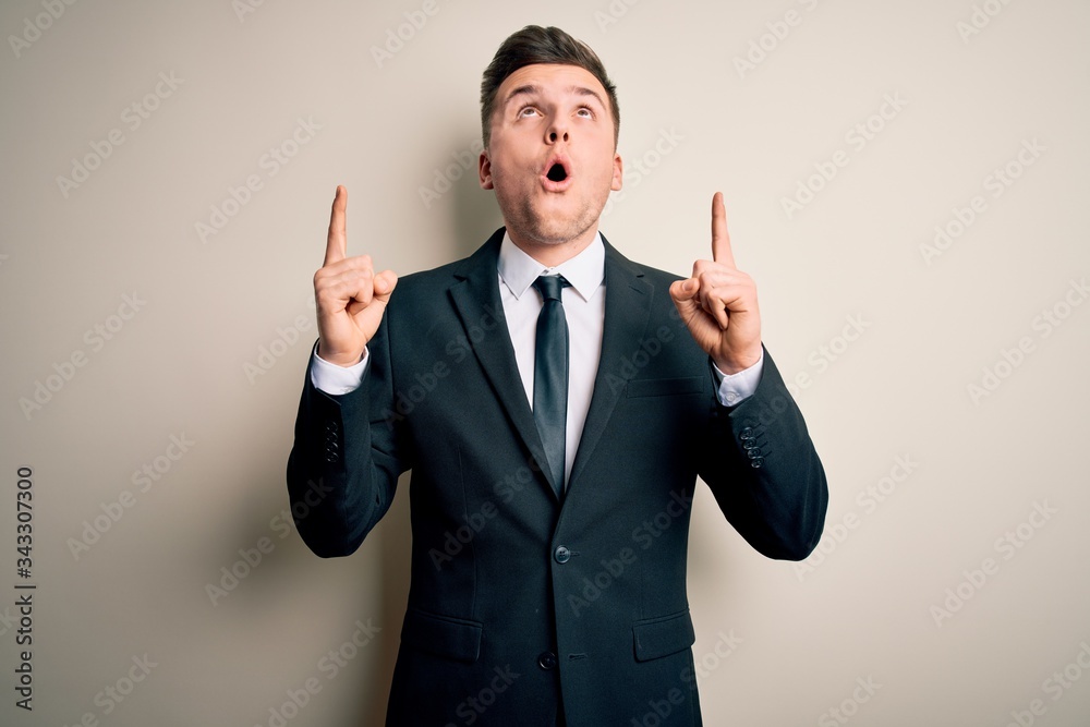 Young handsome business man wearing elegant suit and tie over isolated background amazed and surprised looking up and pointing with fingers and raised arms. Young handsome business man wearing elegant suit and tie over isolated background amazed and surprised looking up and pointing with fingers and raised arms.