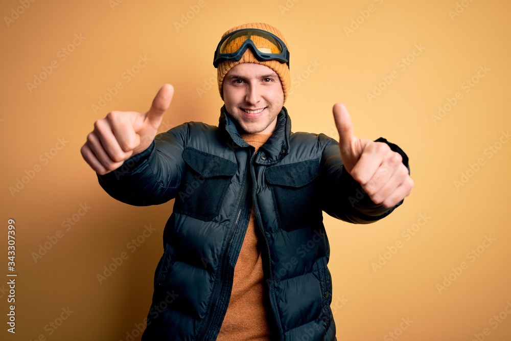Young handsome caucasian man wearing hat, coat and ski glasses for winter and snow weather approving doing positive gesture with hand, thumbs up smiling and happy for success. Winner gesture. Young handsome caucasian man wearing hat, coat and ski glasses for winter and snow weather approving doing positive gesture with hand, thumbs up smiling and happy for success. Winner gesture.